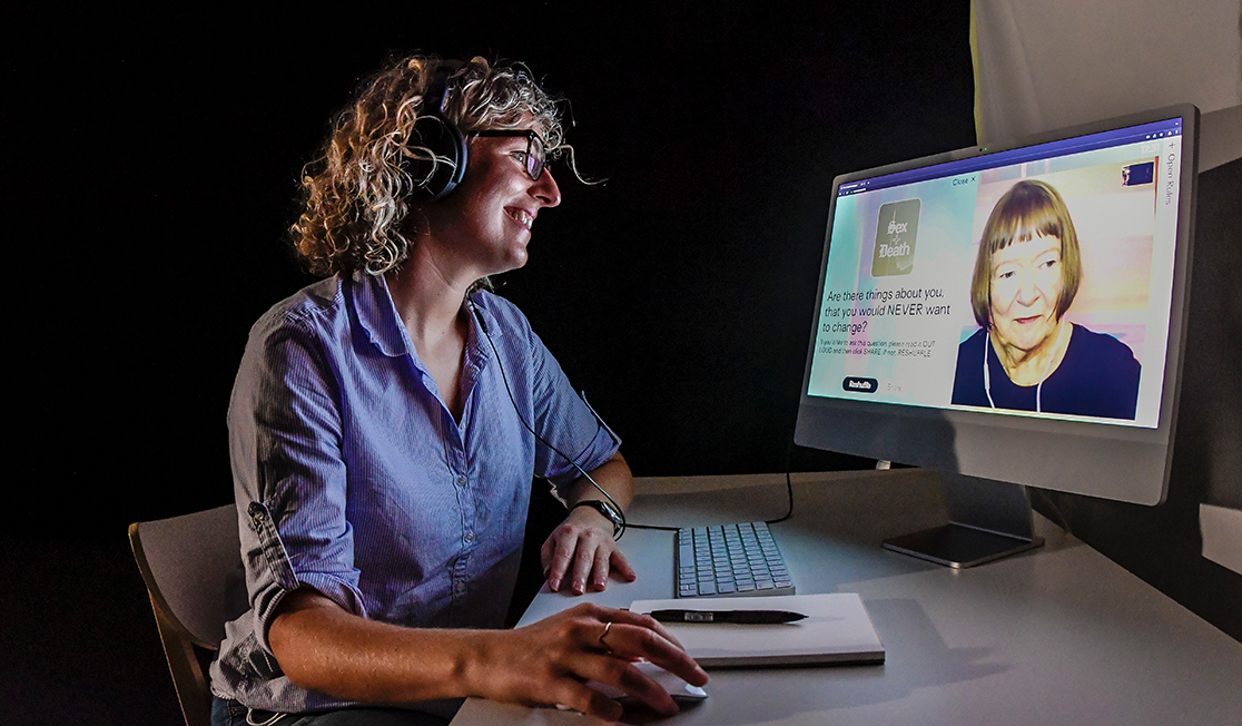 A woman with curly hair and glasses smiles at a computer screen where another women is streaming in