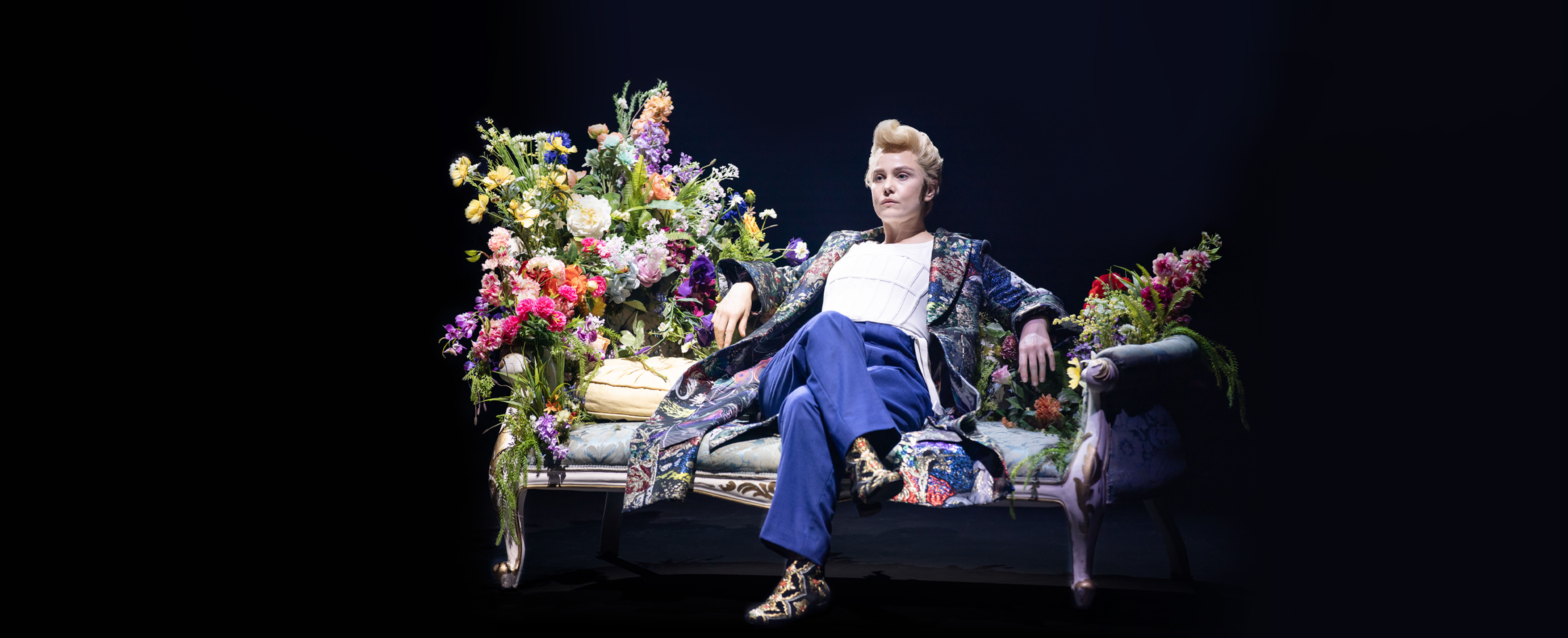 Actor Eryn Jean Norvill sits on a chaise longue with large bouquets of colourful flowers on either side. She is wearing an ornate jacket and boots paired with a white t-shirt and blue trousers.
