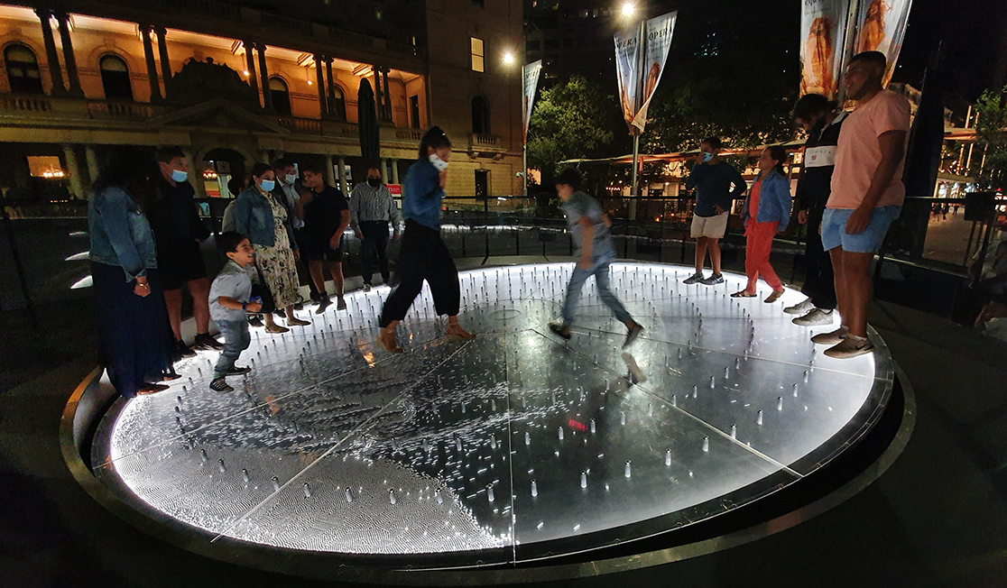 People stand around the edges of a silver disc that's tilted on an angle. Three people are walking through the centre and are the cause of the tilt.