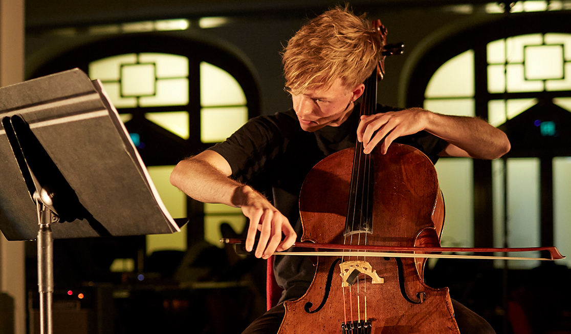 James Morley, a young man with side-parted blond hair and wearing a black t-shirt, is seated and playing the cello.