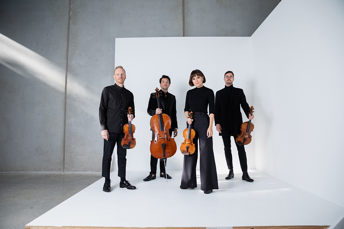 Four musicians dressed in black stand on a white floor with a white wall behind them. They are all holding string instruments.