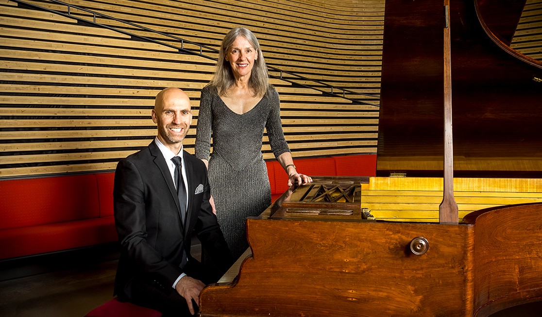 Erin Helyard is wearing a black suit and sitting next to an 1853 Érard piano. Stephanie McCallum stands behind him, wearing a silver dress.