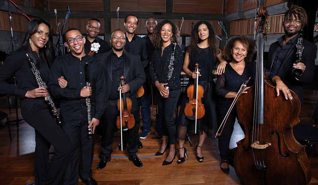 The members of the Chineke! Chamber Ensemble are all wearing black, standing with their instruments and smiling into the camera.