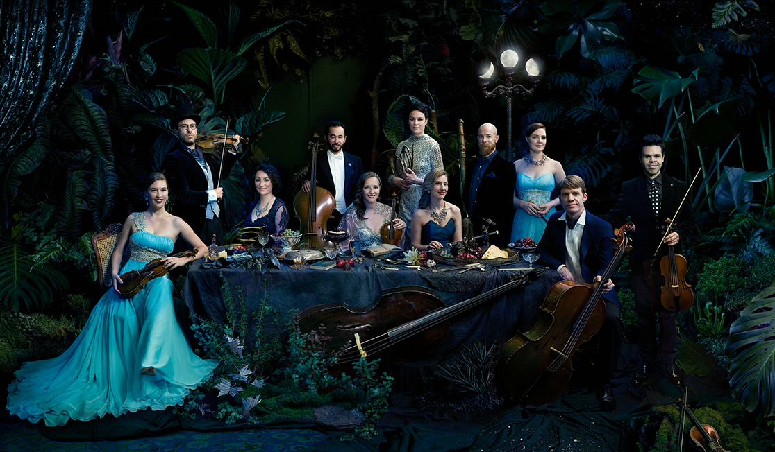 Members of the Australian Haydn Ensemble are seated around a dinner table draped in fabric and laden with fruit in a dark, leafy setting.  They are wearing formalwear and are holding their instruments.