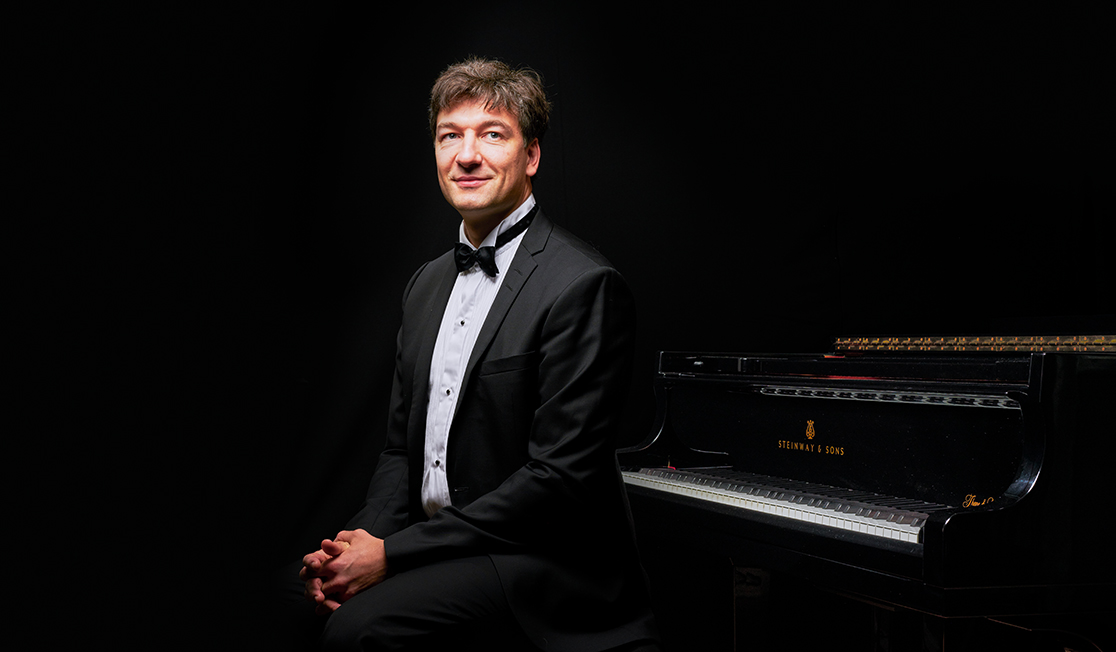 Konstantin Shamray wears a tuxedo and sits next to a piano with his hands clasped in his lap.
