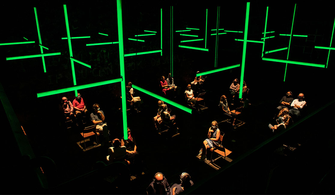 People sit by themselves or in pairs in a darkened room with green fluorescent lights suspended above them.