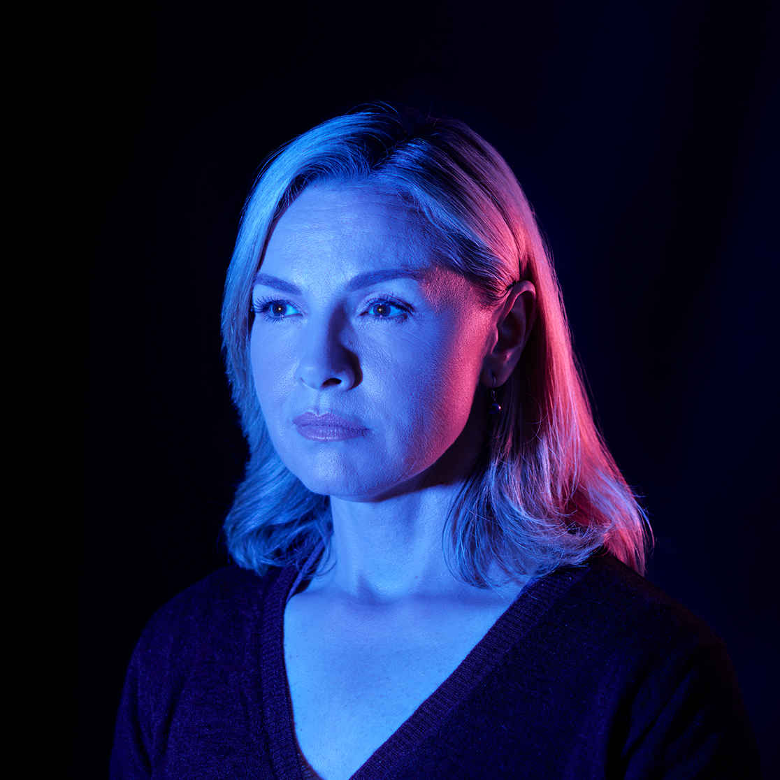 A blonde woman looks to the left of the camera with a serious expression. She is wearing a dark v-neck jumper and is under blue and pink light.