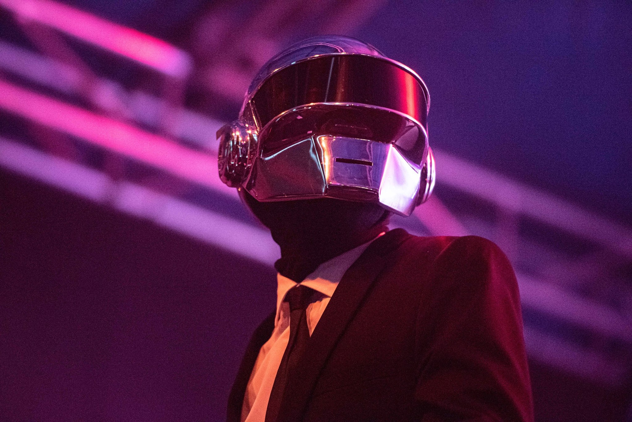 A man dressed in a black suit jacket, white short and black tie has his face hidden within a shiny silver helmet.