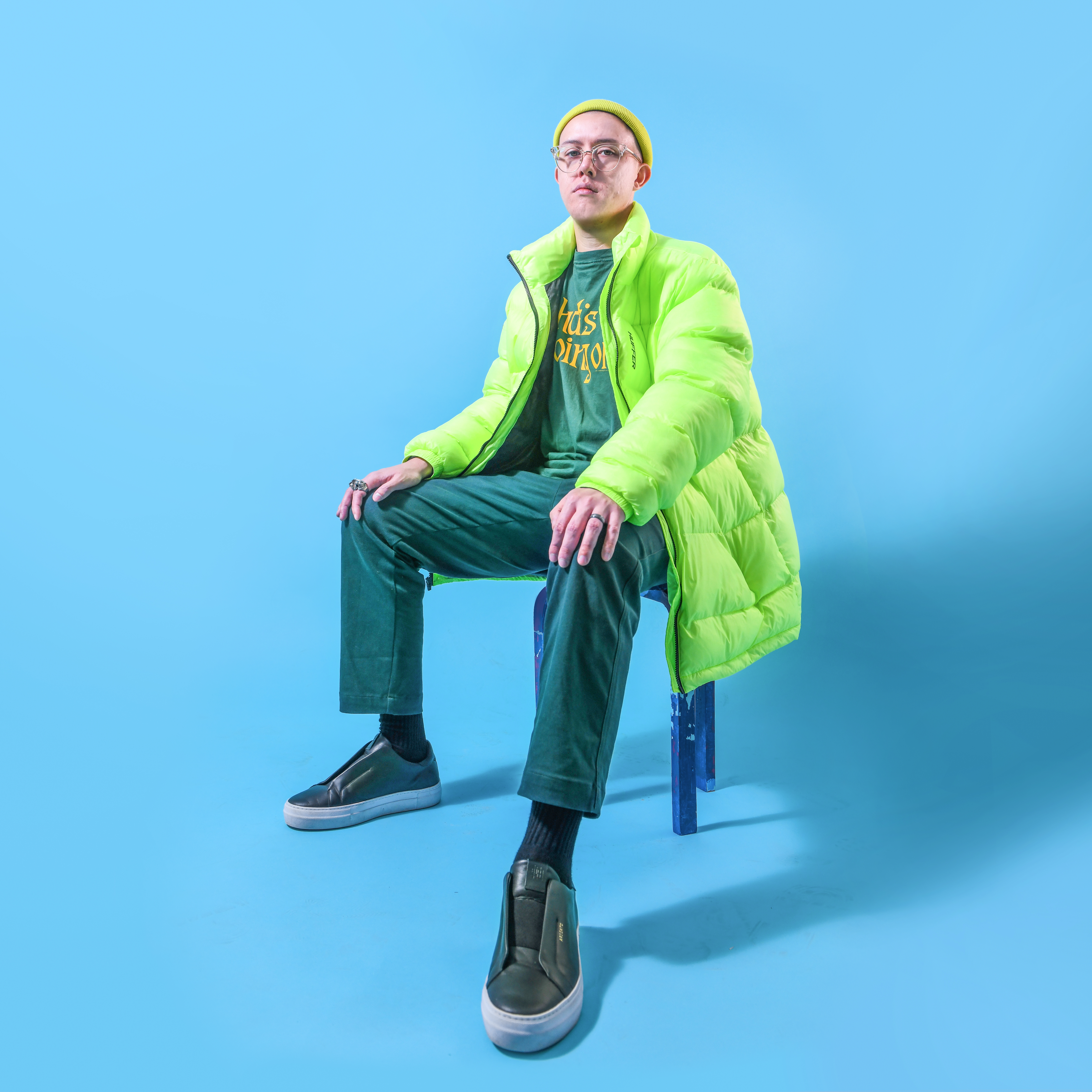 Billy Davis wears a lime green puffer jacket. His t-shirt, pants and shoes are all dark green. He's seated on a dark blue chair in a space where the floor and background are both light blue.