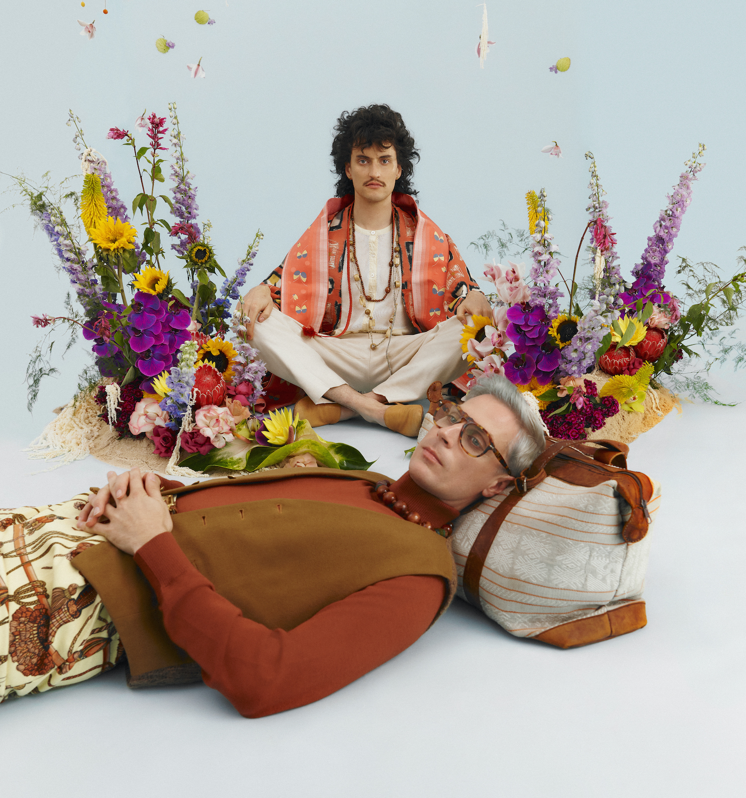 A photo of two men surrounded by flowers and wearing 70s-inspired outfits.