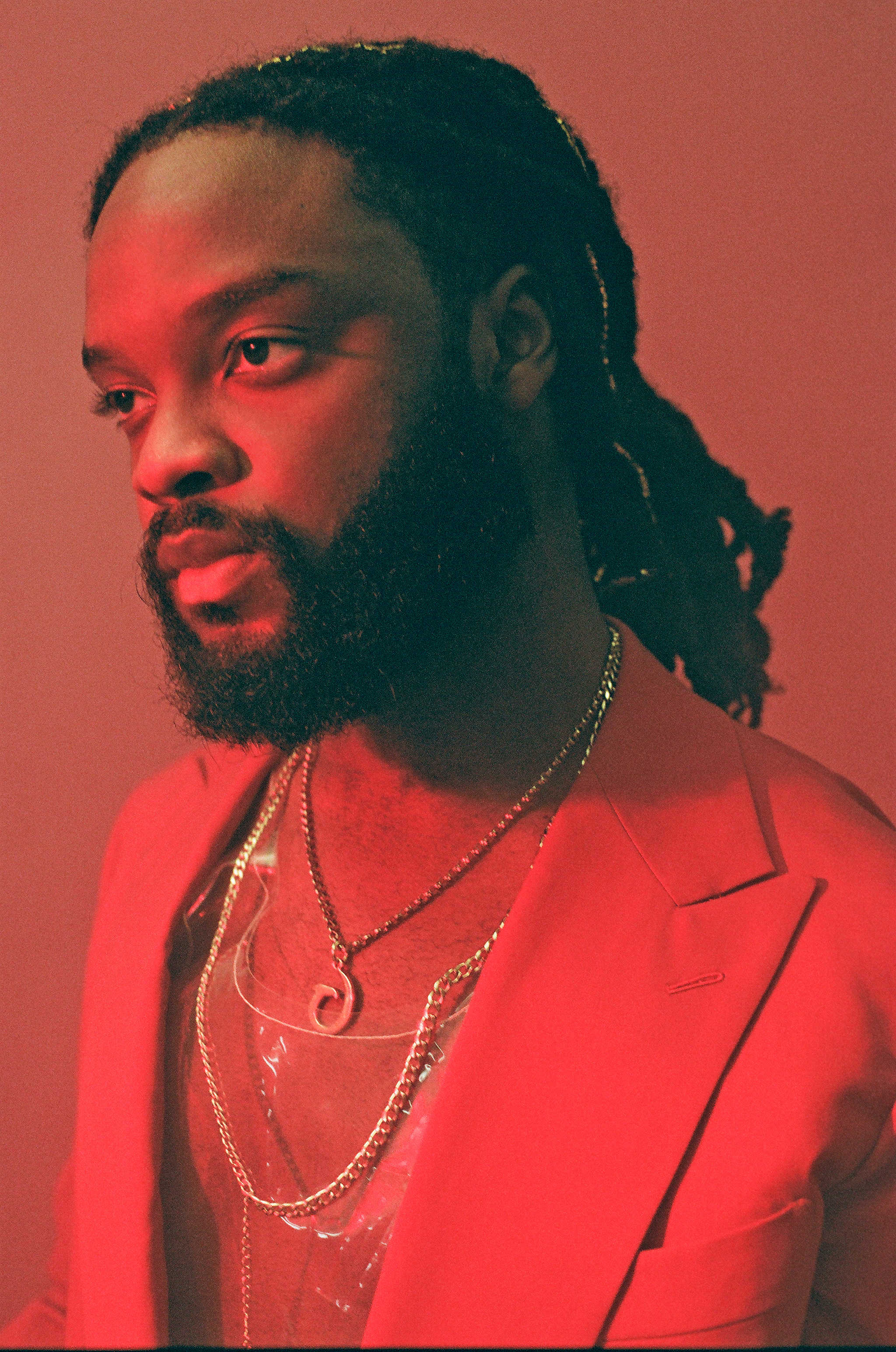 Genesis Owusu, a Ghanian Australian musician with dark dreadlocks and a beard, wears a red blazer and gold chains. Genesis Owusu is looking off to the left and the scene is bathed in red light.