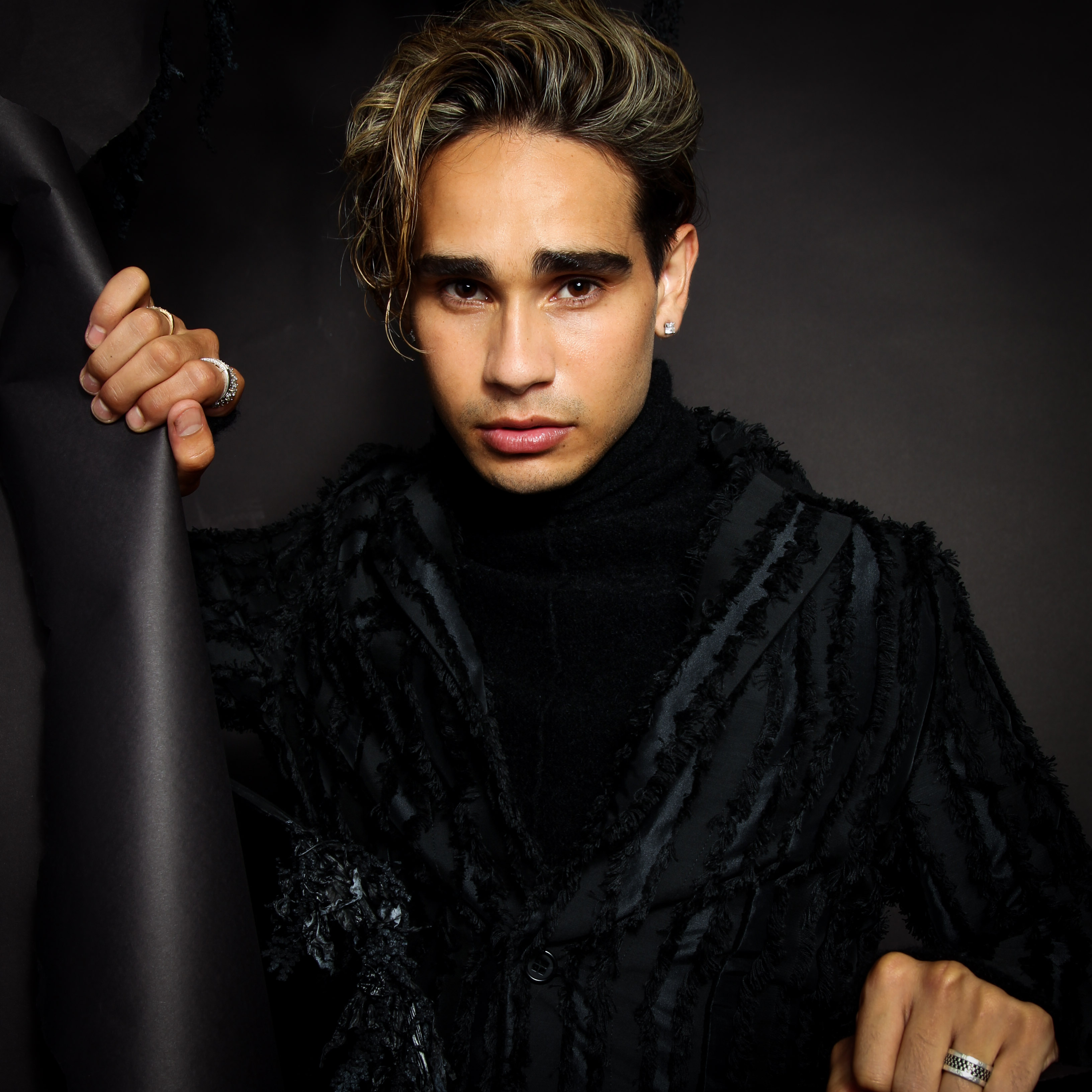 Isaiah Firebrace, a young Aboriginal man with blond-streaked dark hair, wears a black furry coat and looks into the camera with a serious expression. He is holding a piece of black fabric and the background is also black.