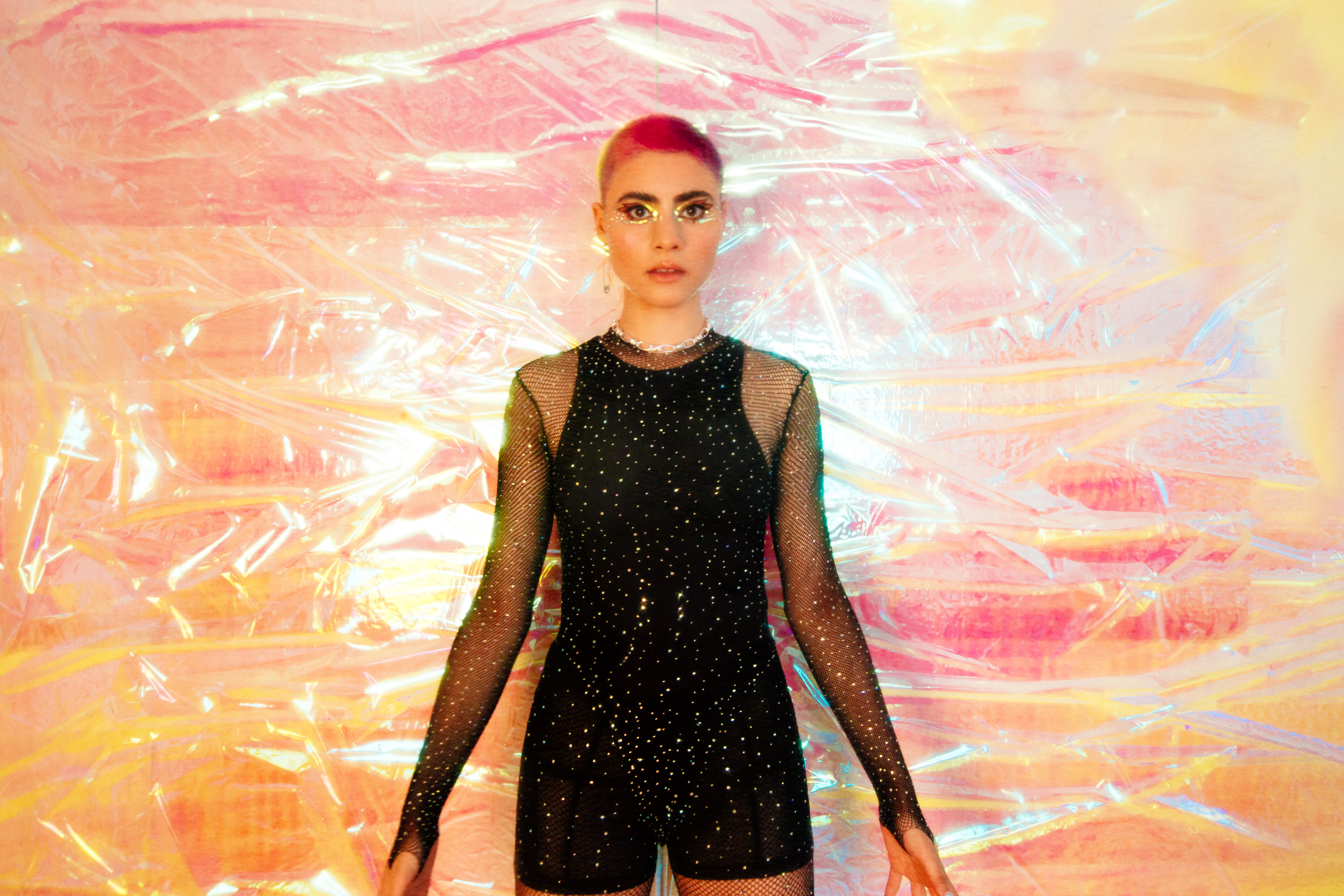Montaigne is a young woman with short hair in a rainbow of bright colours. She wears a sparkly black bodysuit and stares directly into camera with a background that looks like pink and yellow pearlescent cellophane.