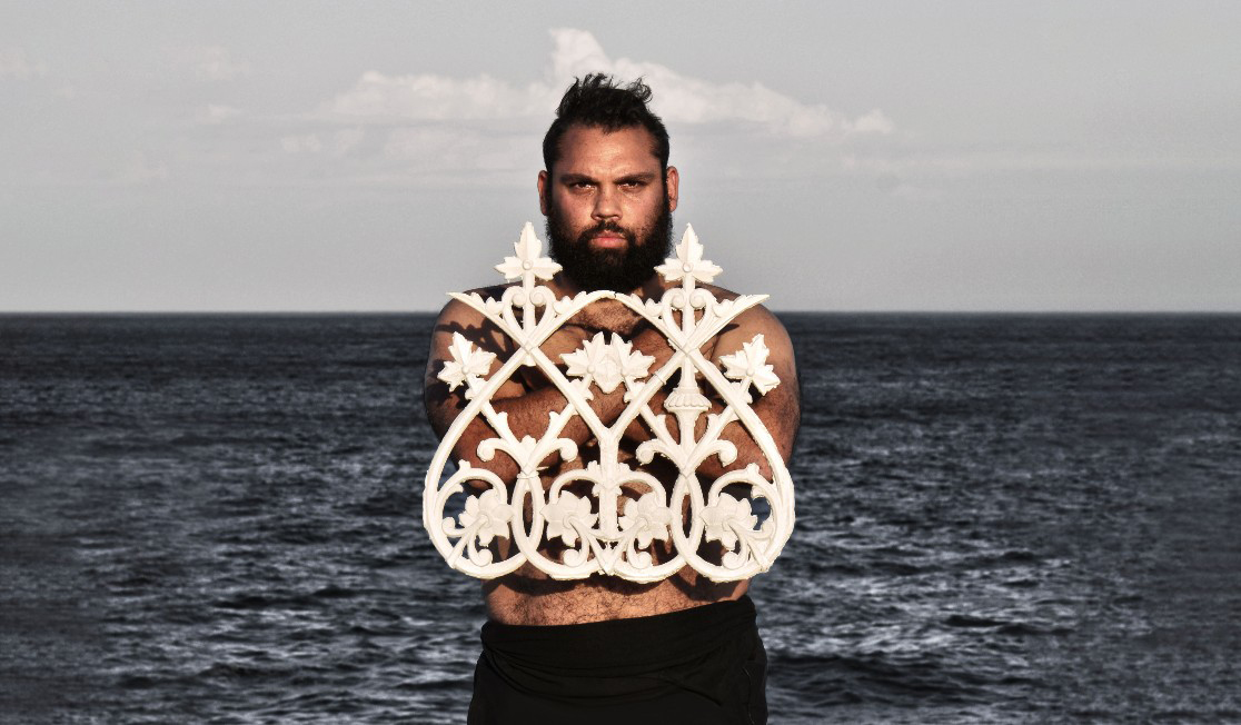 A man with a beard standing in front of the ocean. His arms are crossed on his chest and he is holding a white decorative grate 