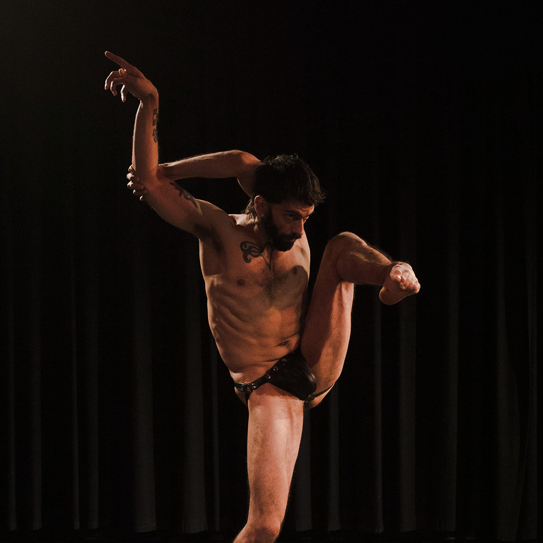 A man in black underwear is pictured on a dark stage. He is balanced on the toes of his right foot, with his left in the air. He has both arms in the air behind his head 
