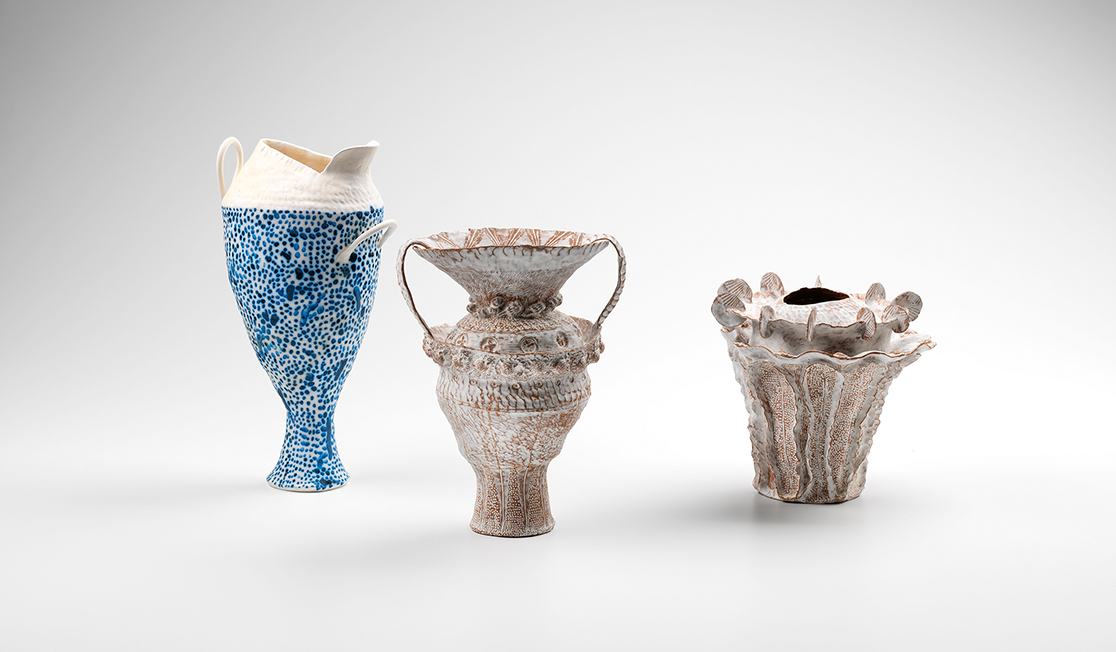 Three handmade terracotta vases on a white backdrop. The left is tall with blue spots and a white top, the middle a terracotta colour with intricate patterns and the right is short and terracotta coloured with white discs around the wavey lip