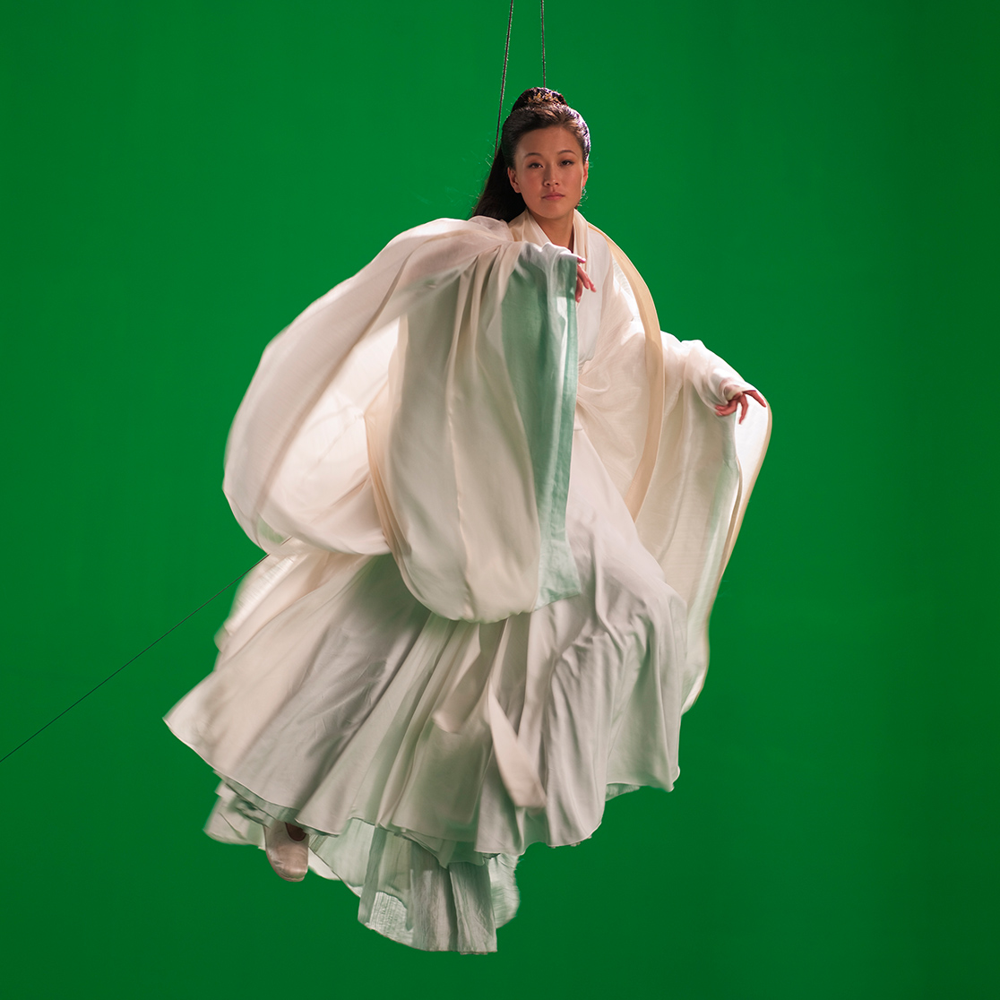 A woman dressed in floaty white fabric is pictured, she is suspended by wires in front of a green screen