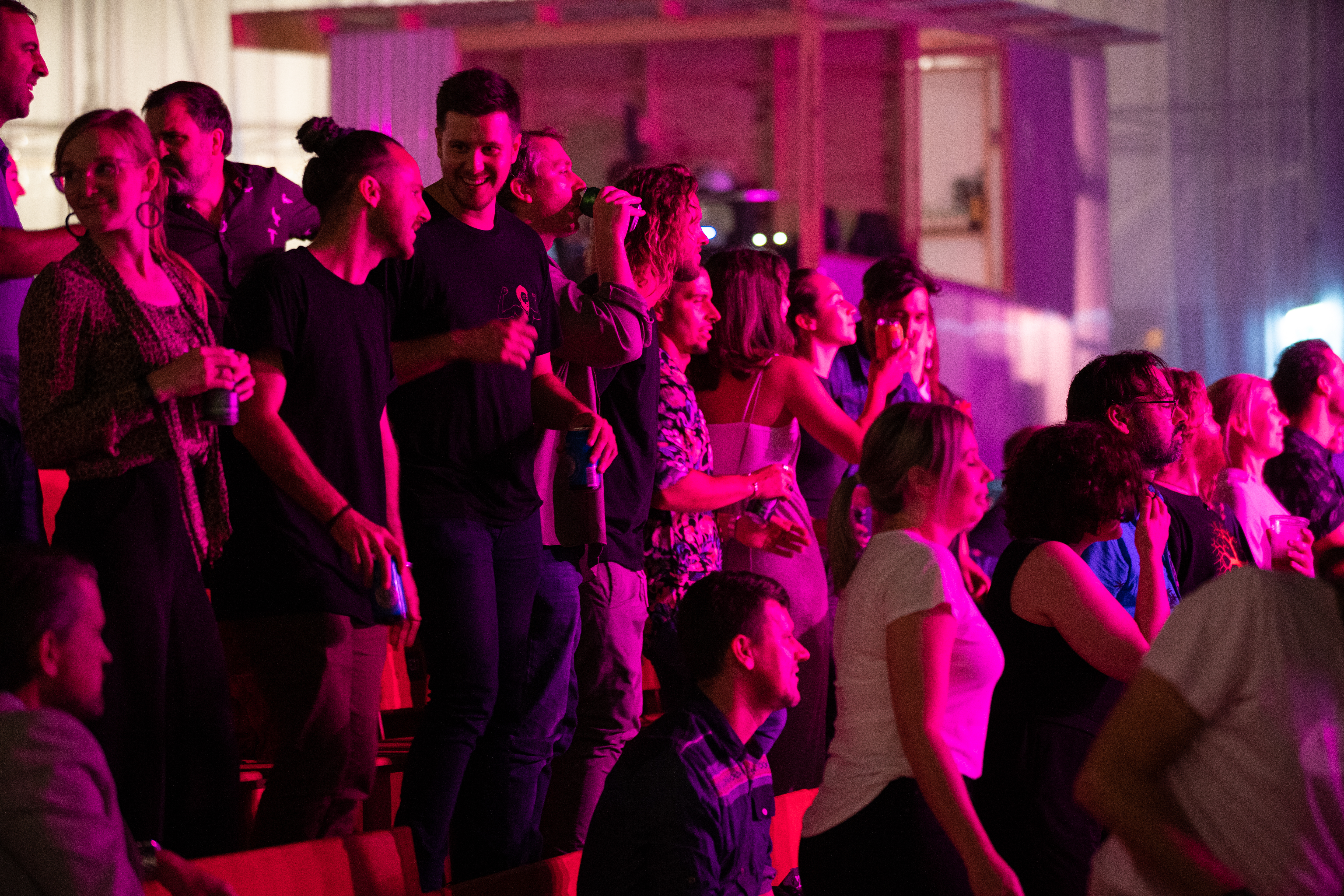 A group of young people watch a performance on stage. They're smiling and bathed in pink light.