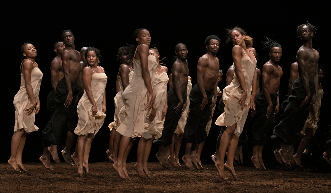A group of dancers dressed in black and beige leap in unison above a dirt floor with a black background.