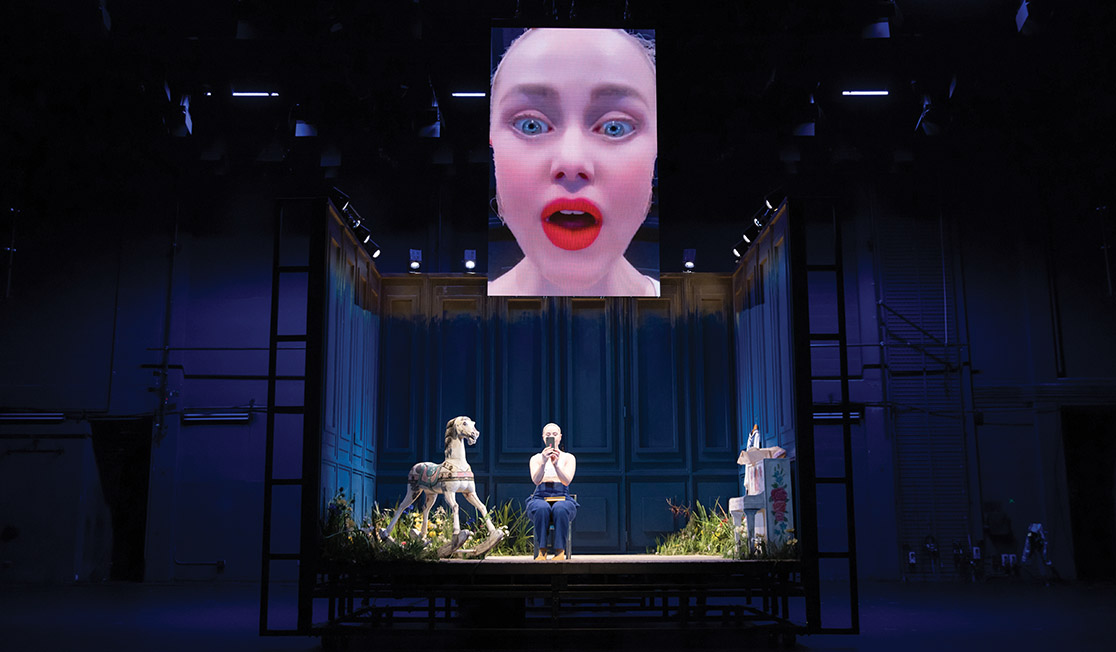 An actor sits in a box on a stage, holding a phone in front of their face. There is a screen above them showing their face (obviously coming from the phone's camera).