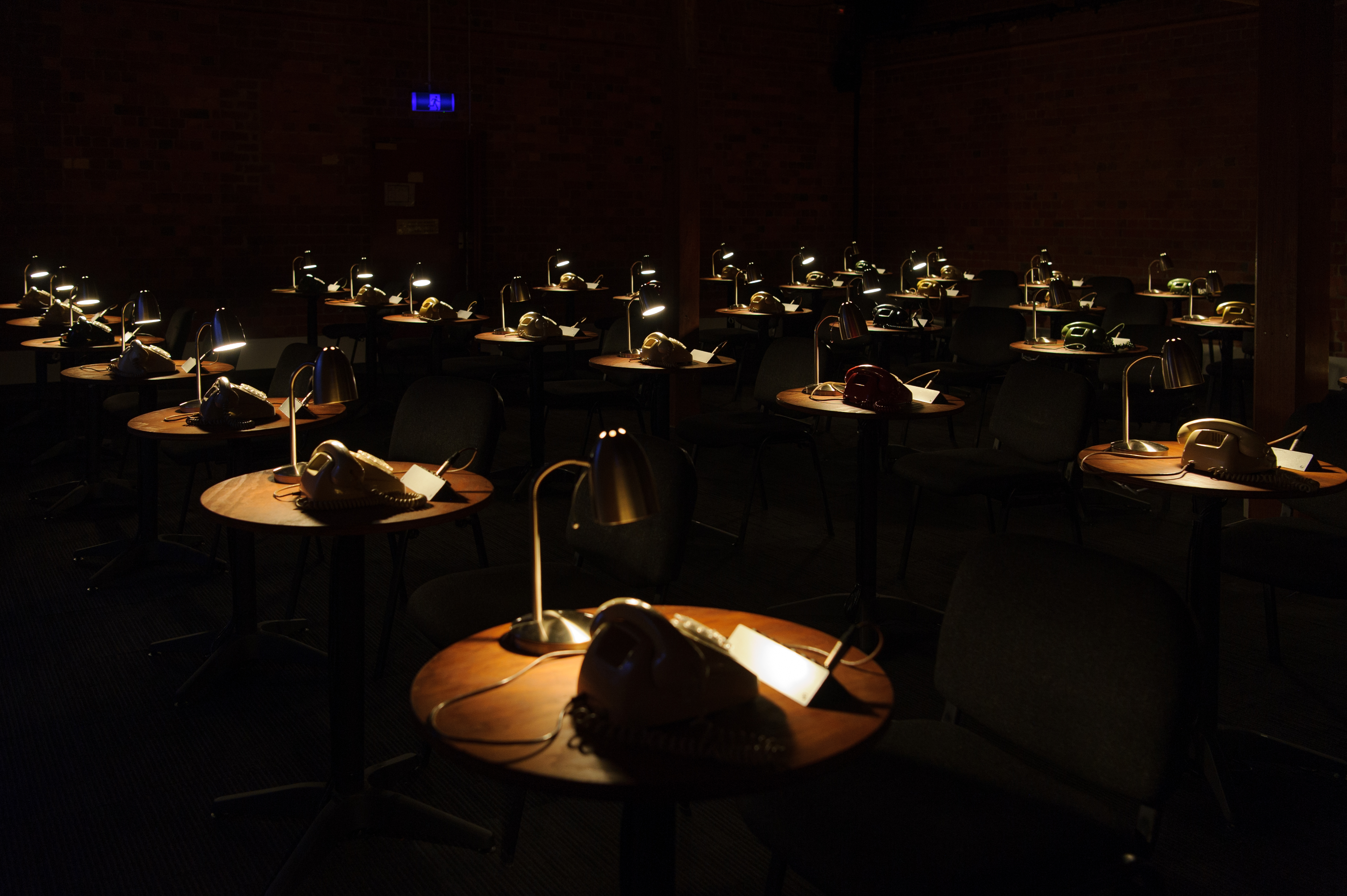 Rows of round desks are pictured in a dark room. On each a rotary dial telephone and a desk lamp.