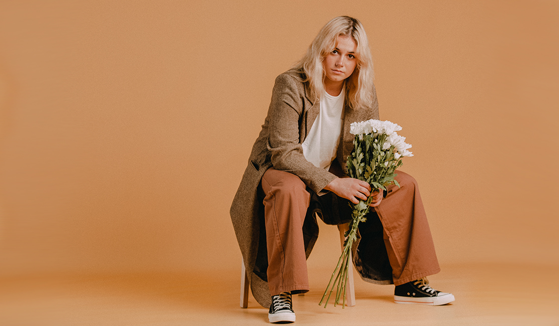 Ruby Fields has blonde hair that falls to just below her shoulders. She is sittng on a chair and is dressed in a beige trenchcoat and rust-coloured pants with black Converse sneakers. She is holding a bunch of white flowers and is looking into the camera with a serious expression.