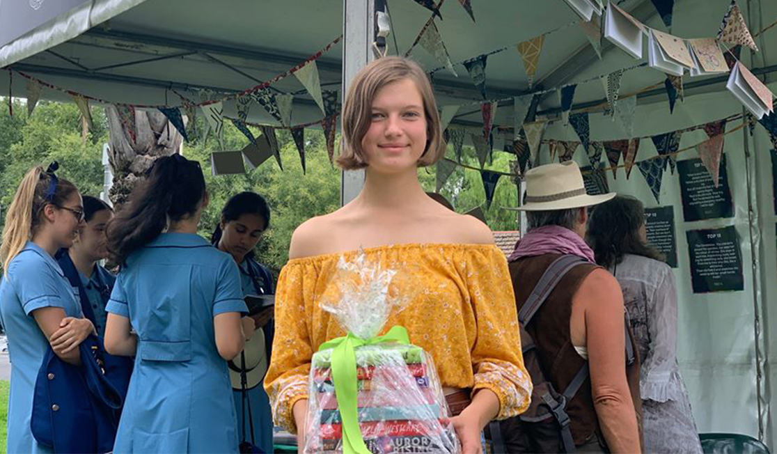 Emily Ashby, the 2020 winner of the microstory competition, has short, light brown hair and is wearing a yellow off-the-shoulder blouse. She is smiling at the camera and holding a stack of books wrapped in cellophane.