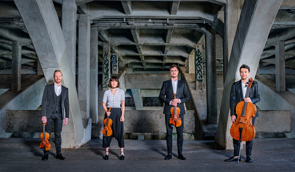 Members of the Australian String Quartet wear black and stand in a row holding their instruments. It appears as though they are standing under a bridge.
