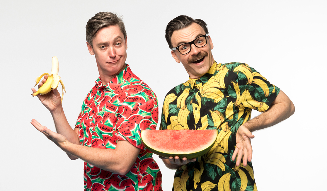 Two men make silly faces at the camera - one is wearing a shirt with watermelon patterns and is holding a banana and the other is wearing a shirt with a banana pattern and holding a sliced watermelon.